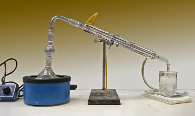 A chemistry lab apparatus.  In the middle thje the Liebig condenser, a pair of long concentric glass tubes, one inside the other, supported by a metal ring stand.  At left a heater heats a flash whose top is connected to the upper end of the inner tube of the condenser.  The condensate collects in a flask at right.  Two rubber tubes connect to the top and bottom of the outer tube, carrying water through it.